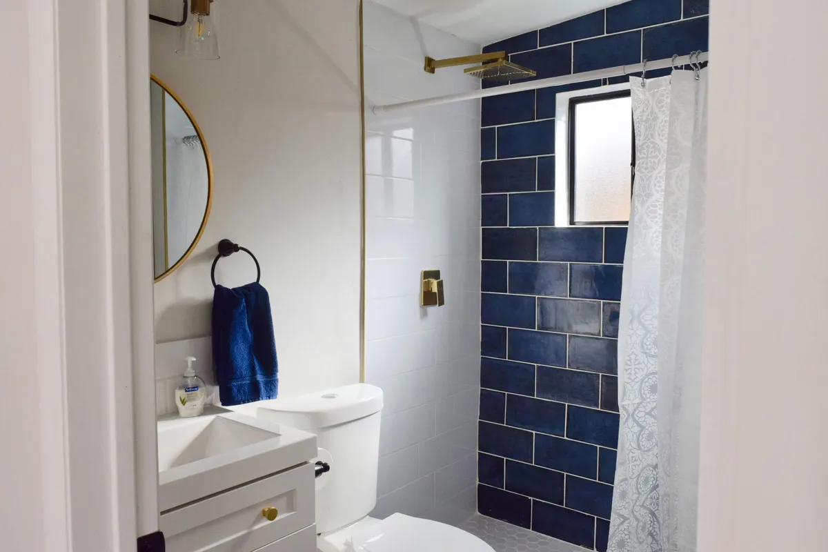 A chic bathroom showcases a blue tiled shower wall, a round mirror above a white sink with gold fixtures. The space feels fresh, bright, and well-appointed.