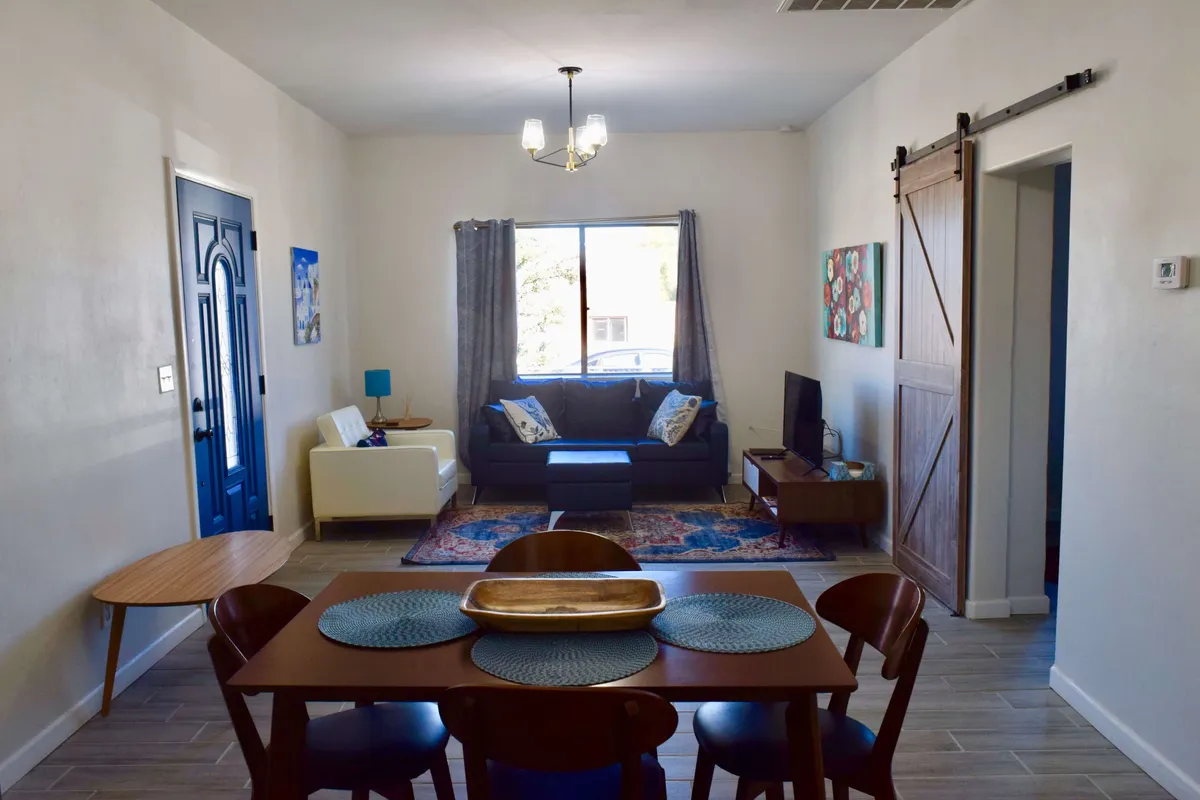 The image showcases a sunlit living and dining space with a striking blue door, a comfortable sofa adorned with colorful decorative pillows, a modern dining table perfect for group meals, and a warm, textured area rug. The space is styled to feel both historic and chic, blending classic adobe charm with inviting, contemporary comfort.