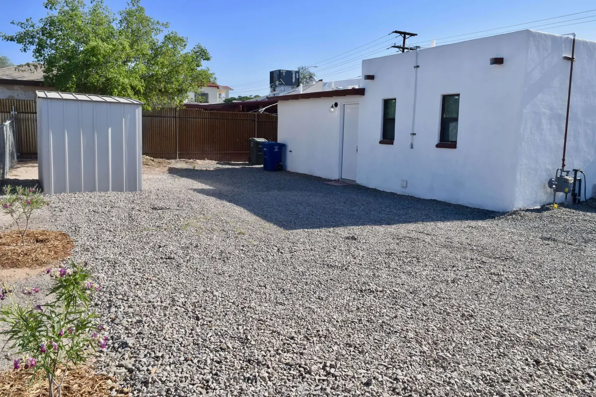 The image features the exterior of a charming historic adobe home, set within a gravel yard accented by a lush green tree. A white building and a practical storage shed stand out as focal points, suggesting both utility and family-friendly outdoor space. The scene hints at activities like gardening, barbecuing, and enjoying sunsets, evoking a welcoming atmosphere perfect for relaxing after exploring Tucson’s attractions.