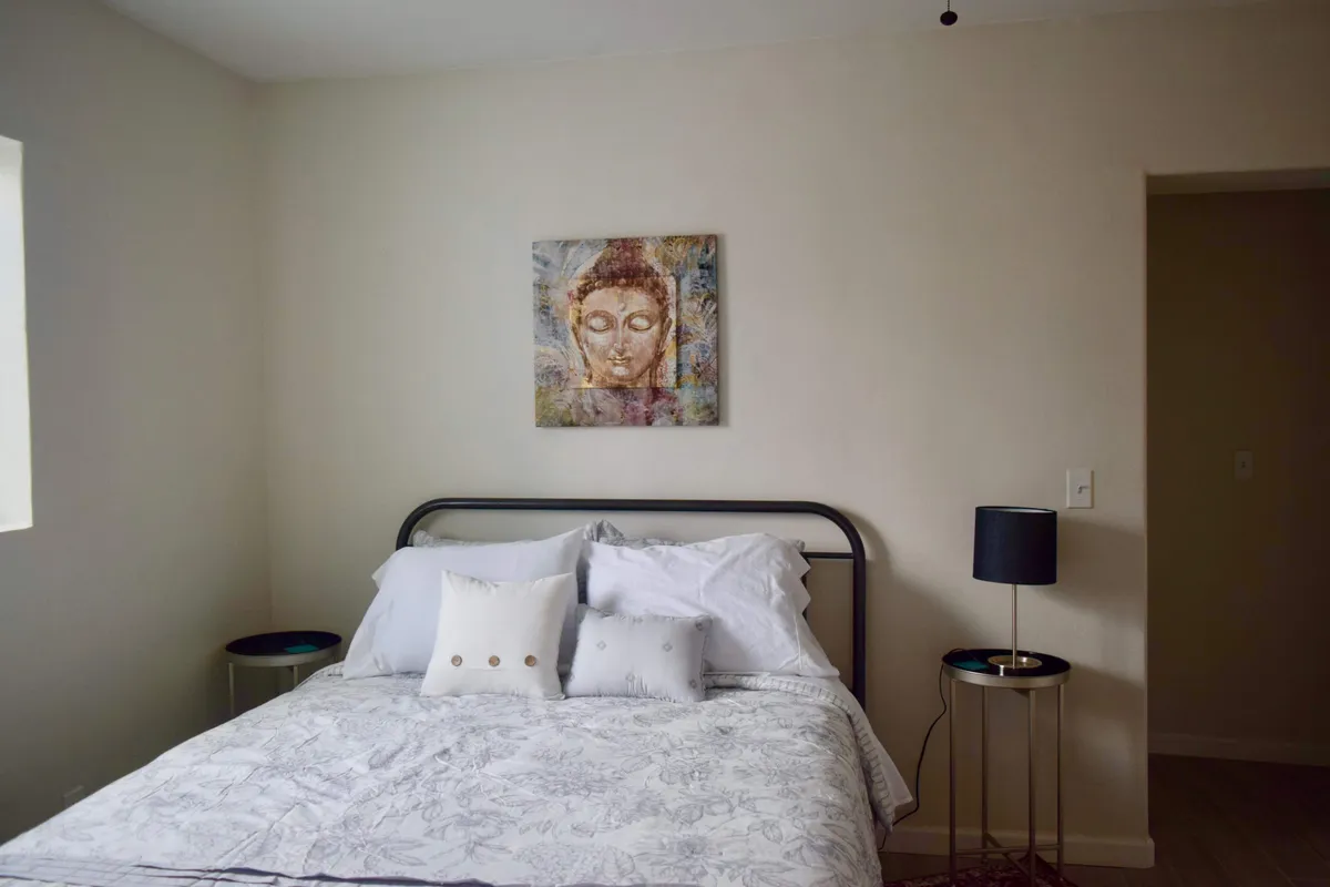 A tranquil bedroom featuring a bed with decorative pillows, serene Buddha artwork above the headboard, and a softly glowing side lamp. The tile flooring and subtle decor create a peaceful atmosphere ideal for relaxation or meditation. The overall tone is inviting and calming—perfect for winding down after a day of adventure.
