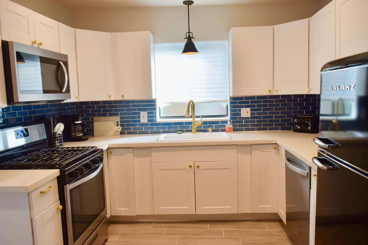 A modern kitchen scene with gleaming stainless steel appliances, crisp white cabinetry, a striking blue tiled backsplash, a golden faucet, and abundant natural light. The open layout flows into a dining area, suggesting a space for meals, socializing, and family gatherings.