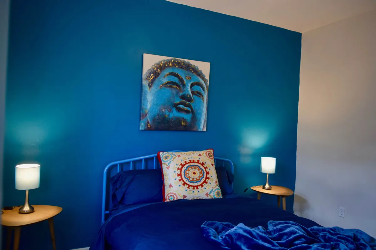 A cozy bedroom with a striking blue accent wall, Buddha artwork, a plush bed with blue bedding and a decorative pillow, and dual table lamps. The room is filled with soft lighting and tranquil energy, inviting restful sleep, quiet reading, or meditative mornings.