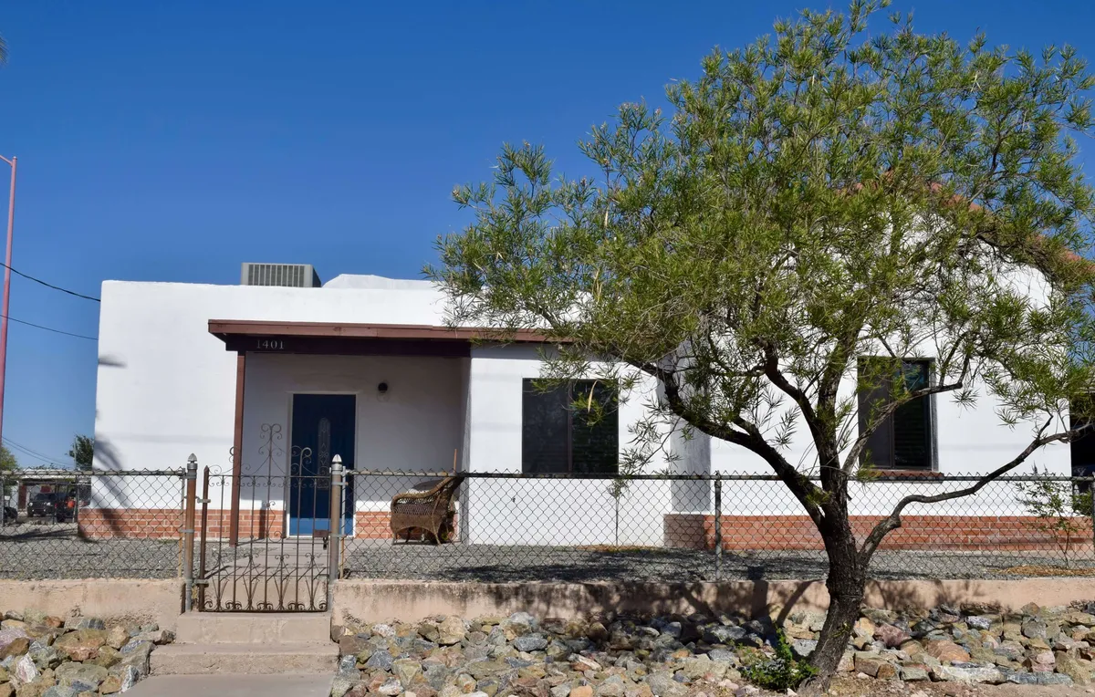 This image displays the home's front entrance, complete with a decorative fence and a mature tree in the yard. The area is tidy and welcoming, with a hint of the vibrant neighborhood visible beyond the gate. The entrance area has character and a sense of authentic Tucson charm, suggesting easy access for arrivals and departures.